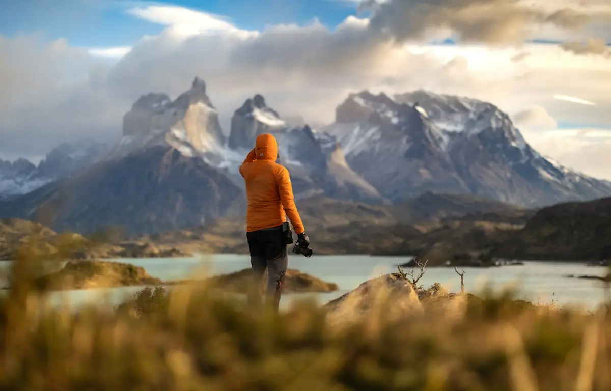 Pessoa de jaqueta laranja diante de montanhas nevadas e um lago
